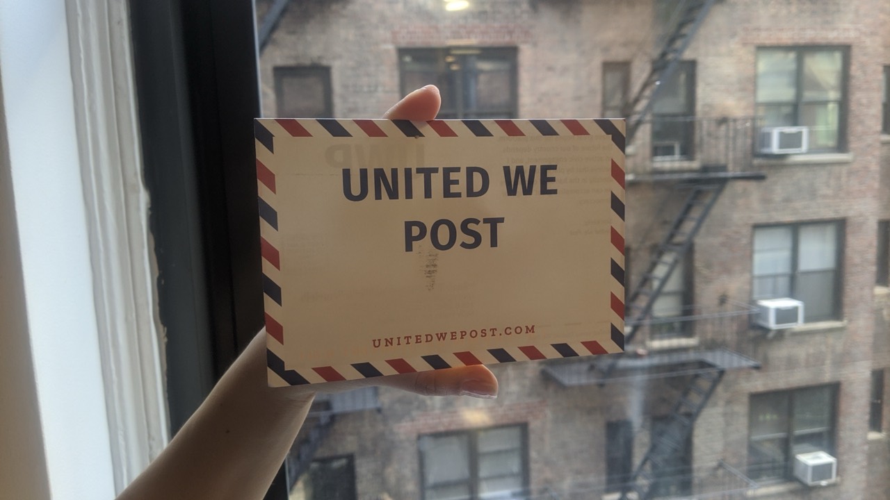 A hand holds a United We Post postcard in front of a city apartment window.